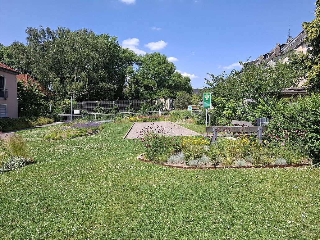 Für Anwohner*innen nutzbare Grünfläche inmitten von Wohnbebauung mit Blumenstreifen und Bouleplatz