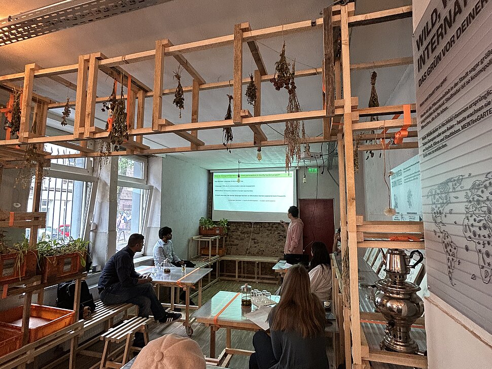 Innenraum der Pop-Up-IGA Duisburg während eines Seminars der TU Dortmund. Mehrere Studierende sitzen an Tischen und schauen auf eine Leinwand, auf die etwas mit einem Beamer projiziert wird. 