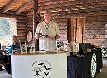 Vesperto Fledermausschutz_Duisburg_Tom Perkowski_2025 In einer rustikalen Holzhütte (Duisburger Zoo) steht ein Mann hinter einem halbhohen, gebogenen Infostand. Auf der Front ist das Logo „VESPERTO Fledermausschutz Niederrhein / Rhein-Ruhr“ zu sehen. Der Mann trägt ein helles Hemd, eine Kappe und ein Namensschild und hält ein kleines Fledermausmodell in den Händen.