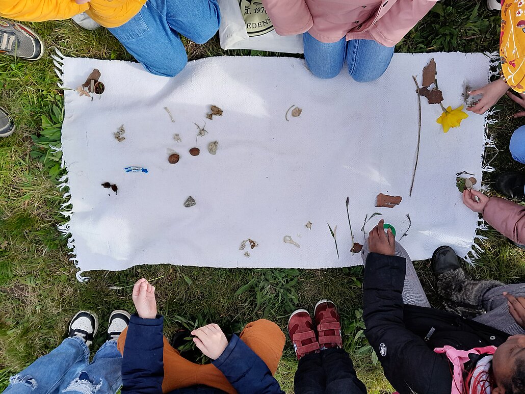 Zu sehen ist ein weißes Tuch mit unterschiedlichen Naturgegenständen: Blättern, Pflanzenteilen und auch Knochen. Das Foto wurde von oben nach unten aufgenommen und zu sehen sind auch Ausschnitte von Füßen, Händen und Schuhen von Kindern, die an dem Tuch kien.