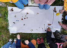 Zu sehen ist ein weißes Tuch mit unterschiedlichen Naturgegenständen: Blättern, Pflanzenteilen und auch Knochen. Das Foto wurde von oben nach unten aufgenommen und zu sehen sind auch Ausschnitte von Füßen, Händen und Schuhen von Kindern, die an dem Tuch kien.