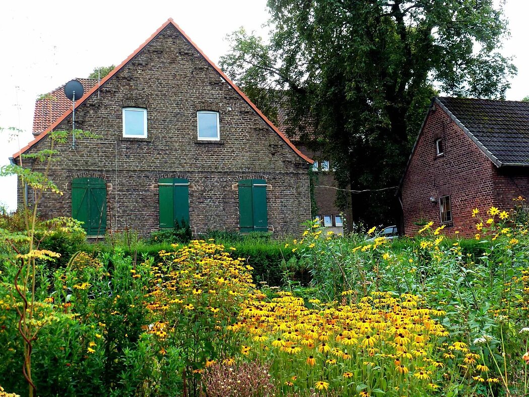 Privatgarten mit Stauden im Vordergrund und einer Hofanlage im Hintergrund