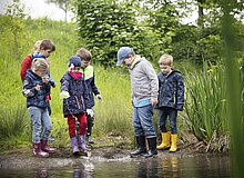 Zu sehen sind Kinder, die mit Regenklamotten und Gummistiefeln durch eine Pfütze im Grünen waten und sich diese anschauen