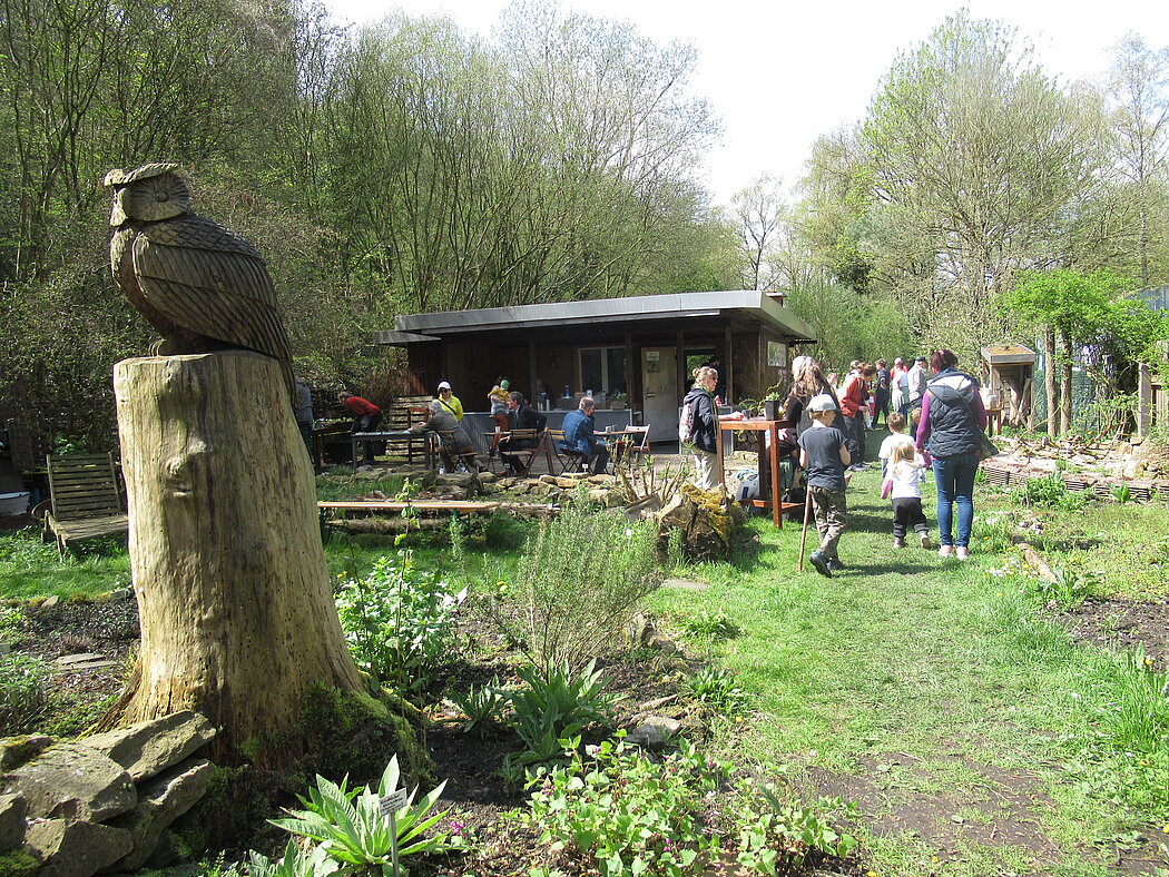 Naturgarten mit Pflanzbeeten, Laube, Eulenskulptur und Besuchern