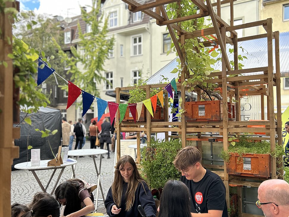 Außenbereich der Pop-Up-IGA Duisburg während des Straßenfests. Mehrere Jugendliche sitzen an einem Tisch und basteln. 