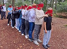 Das Foto zeigt ein lichtes Waldstück, das unterholzfrei ist. Eine Gruppe von Jugendlichen steht mit verundenen Augen hiterinander und fasst s