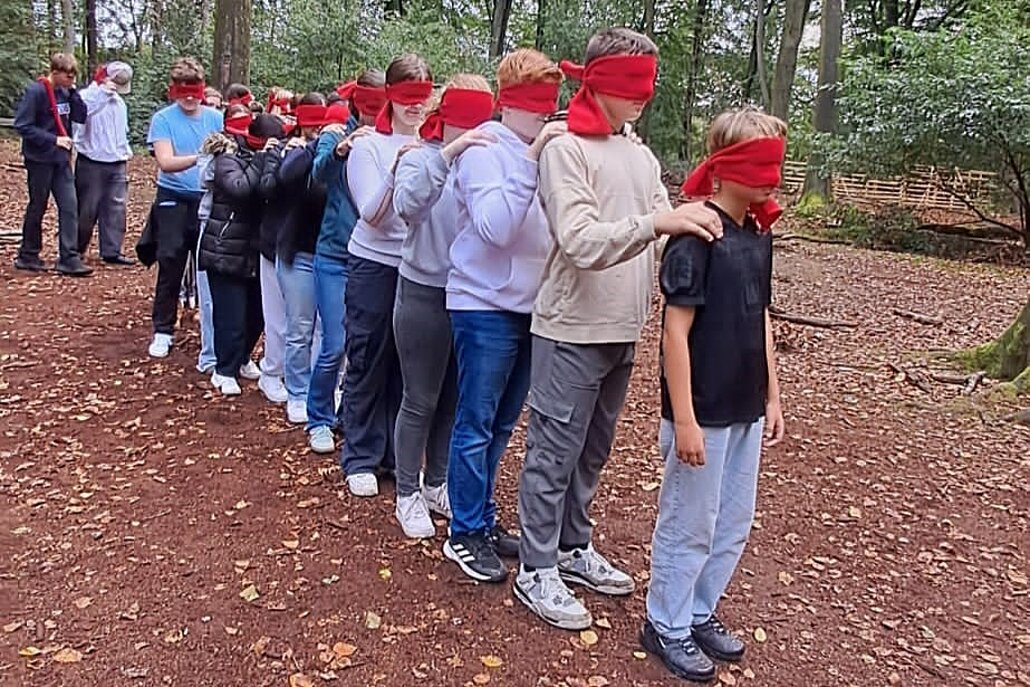 Das Foto zeigt ein lichtes Waldstück, das unterholzfrei ist. Eine Gruppe von Jugendlichen steht mit verundenen Augen hiterinander und fasst s