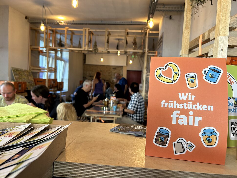 Innenraum der Pop-Up-IGA Duisburg. Im Vordergrund ein orangener Aufsteller auf dem Tresen mit der Aufschrift "Wir frühstücken fair". Im Hintergrund Menschen sitzend an Tischen beim essen.