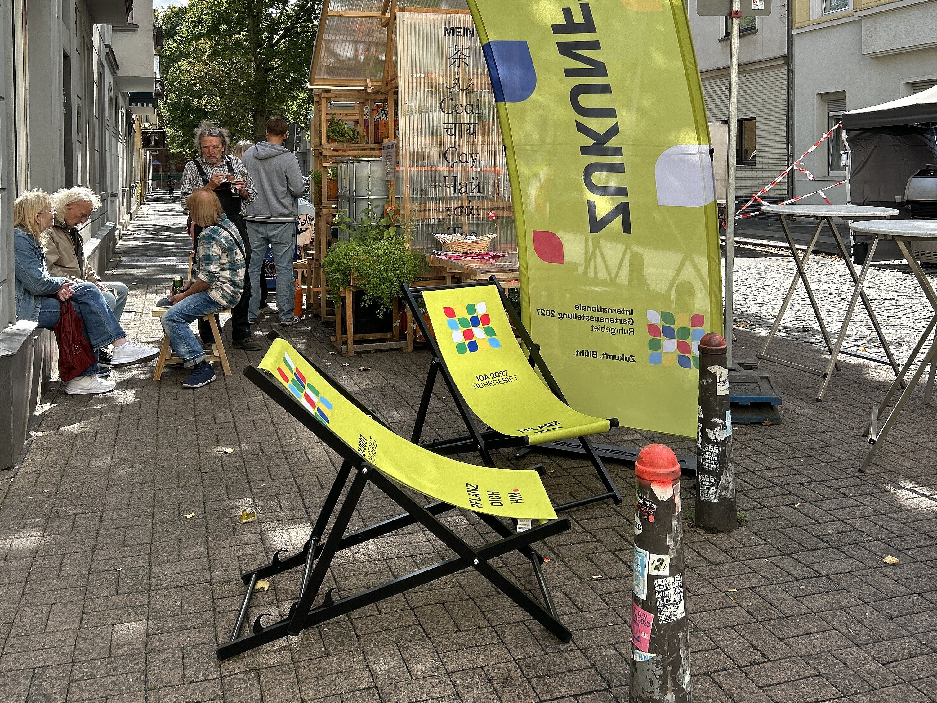 Außenbereich der Pop-Up-IGA Duisburg. Zwei grüne Liegestühle mit dem IGA-Logo überspannt von einem grünen Bannerbow mit IGA-Logo. Im Hintergrung sitzende Mesnchen, die sich unterhalten.