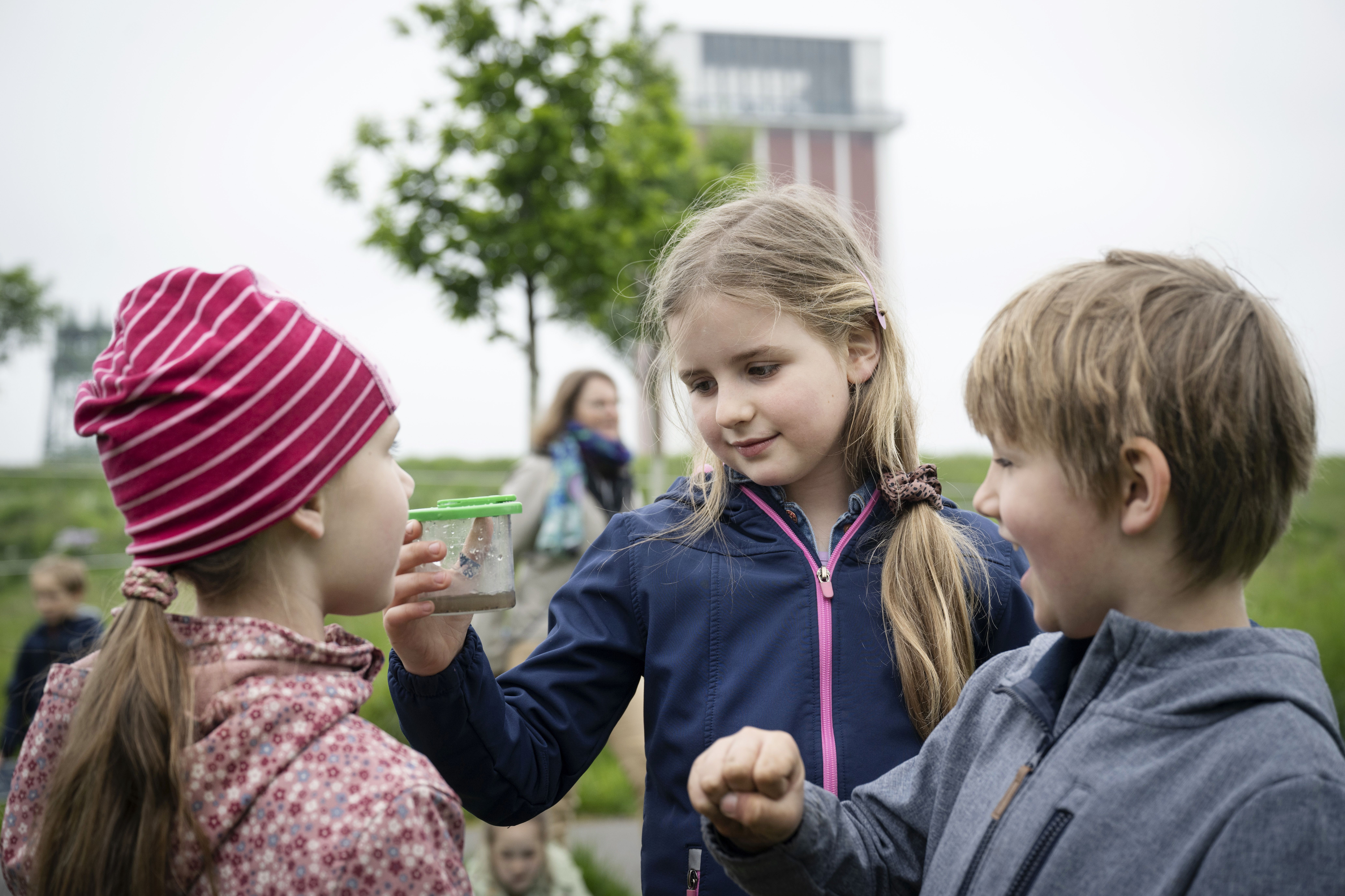 Zu sehen sind drei Grundschulkinder, die nebeneinander stehen und gemeinsam auf eine Becherlupe schauen, die eines der Kinder hält. Im unscharfen Hintergrund ist der rostrote Förderturm der alten Zeche Friedrich-Heinrich zu sehen sowie ein Baum.